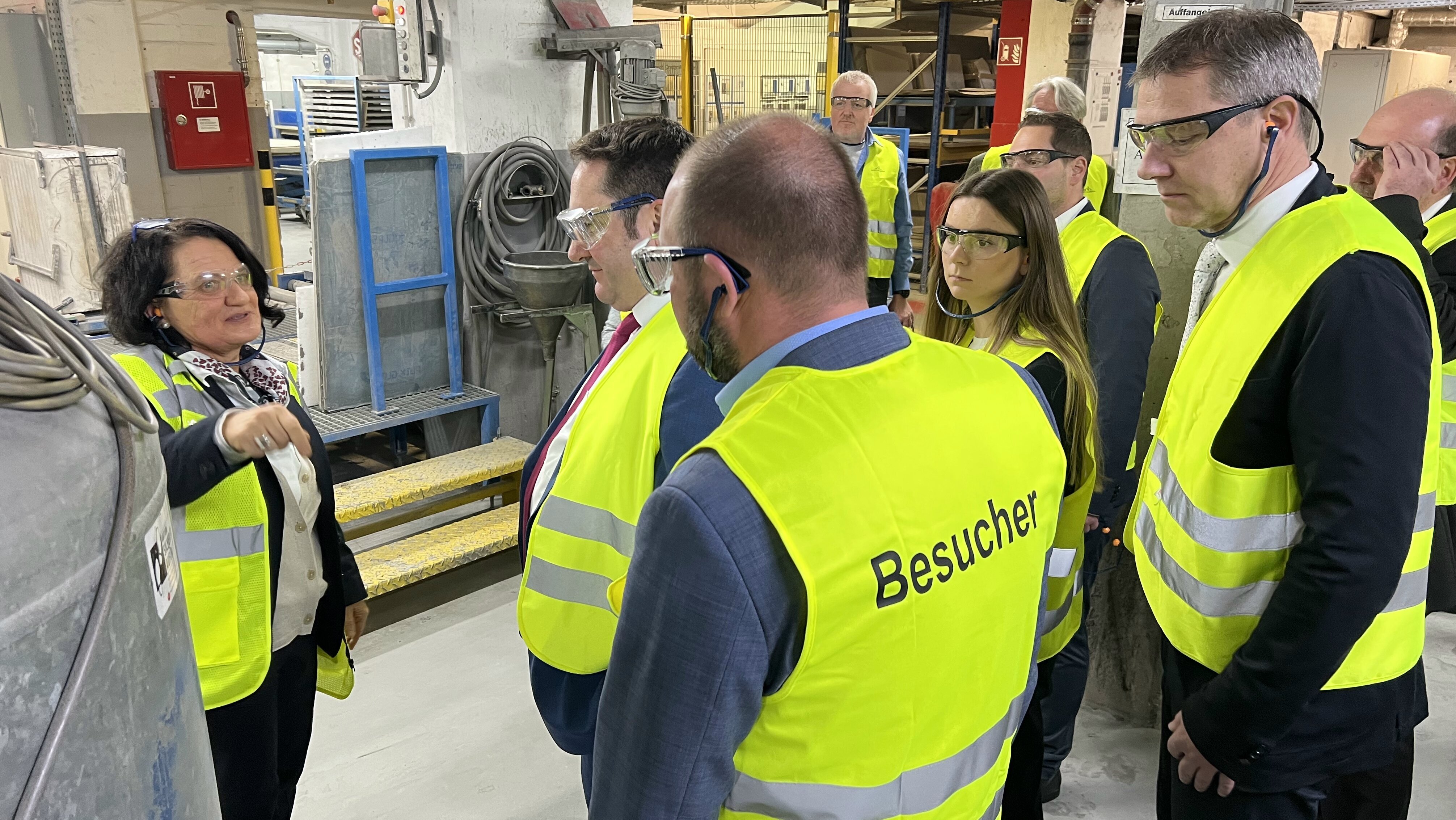 Eine Personengruppe bekleidet mit gelben Sicherheitswesten steht in einem Raum mit Produktionsmaschinen, die man im Hintergrund sieht. In der Mitte steht Geschäftsführerin Brigtte Wirth, die etwas erklärt.