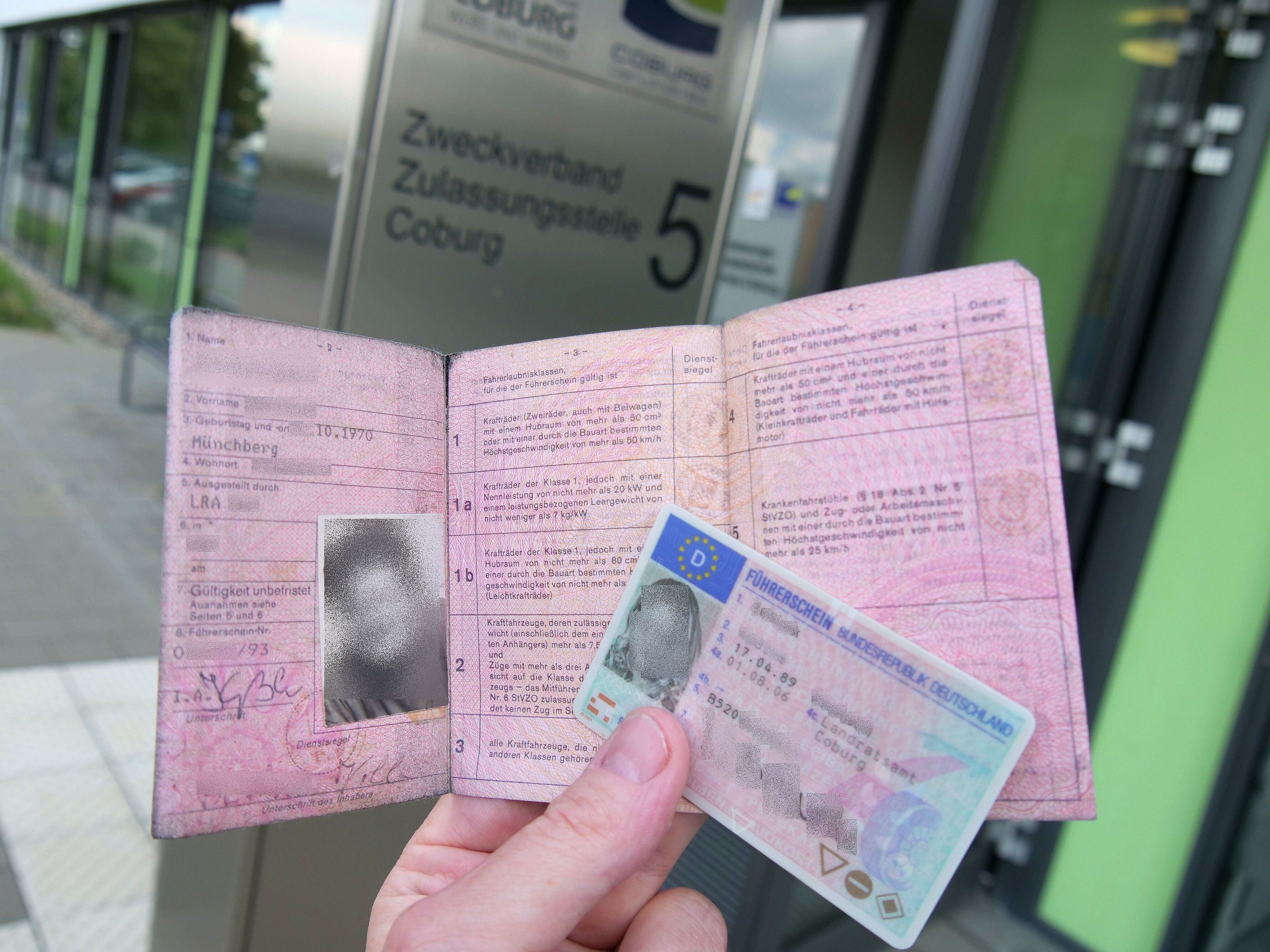 Eine Hand hält einen alten rosafarbenen sowie einen neuen Kartenführerschein. Im Hintergrund ist der Eingang zur Zulassungsstelle Coburg zu sehen.