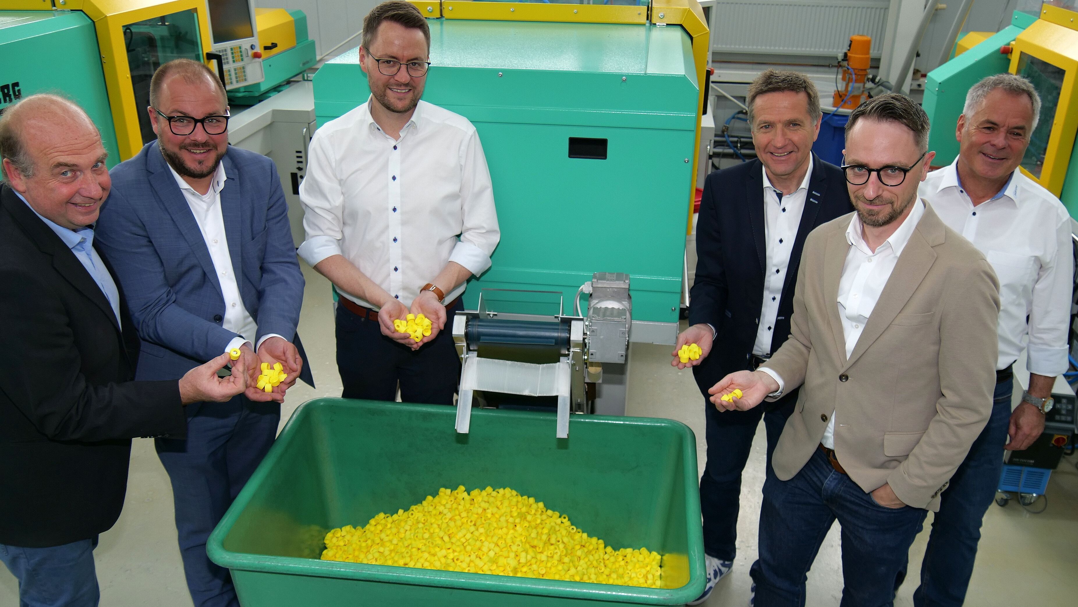 Martin Schmitz, Sebastian Straubel, Dr. Sebastian Heim, Karl Kolb, Cedric Lindner und Matthias Heim stehen vor einer grünen Maschine, aus der kleine, gelbe Kunststoff-Verschlüsse für Flaschen fallen.