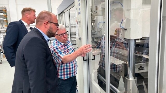 Matthias Bohl, Sebastian Straubel und Michael Keilich blicken durch eine Glasscheibe auf das Innere einer Maschine, in der Kunststoffteile verarbeitet werden.