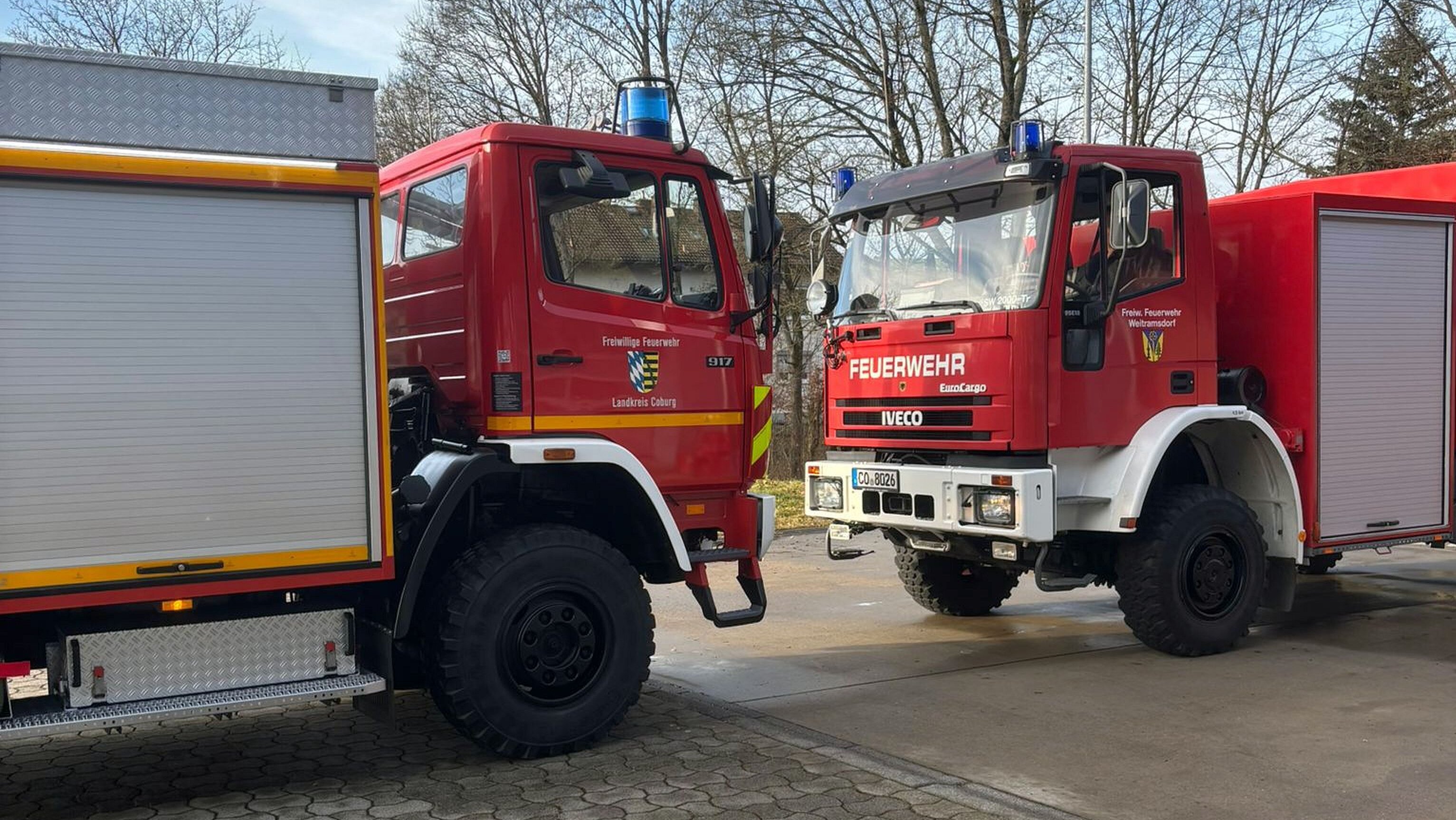 Zwei rote Feuerwehrfahrzeuge stehen auf einer betonierten Fläche.
